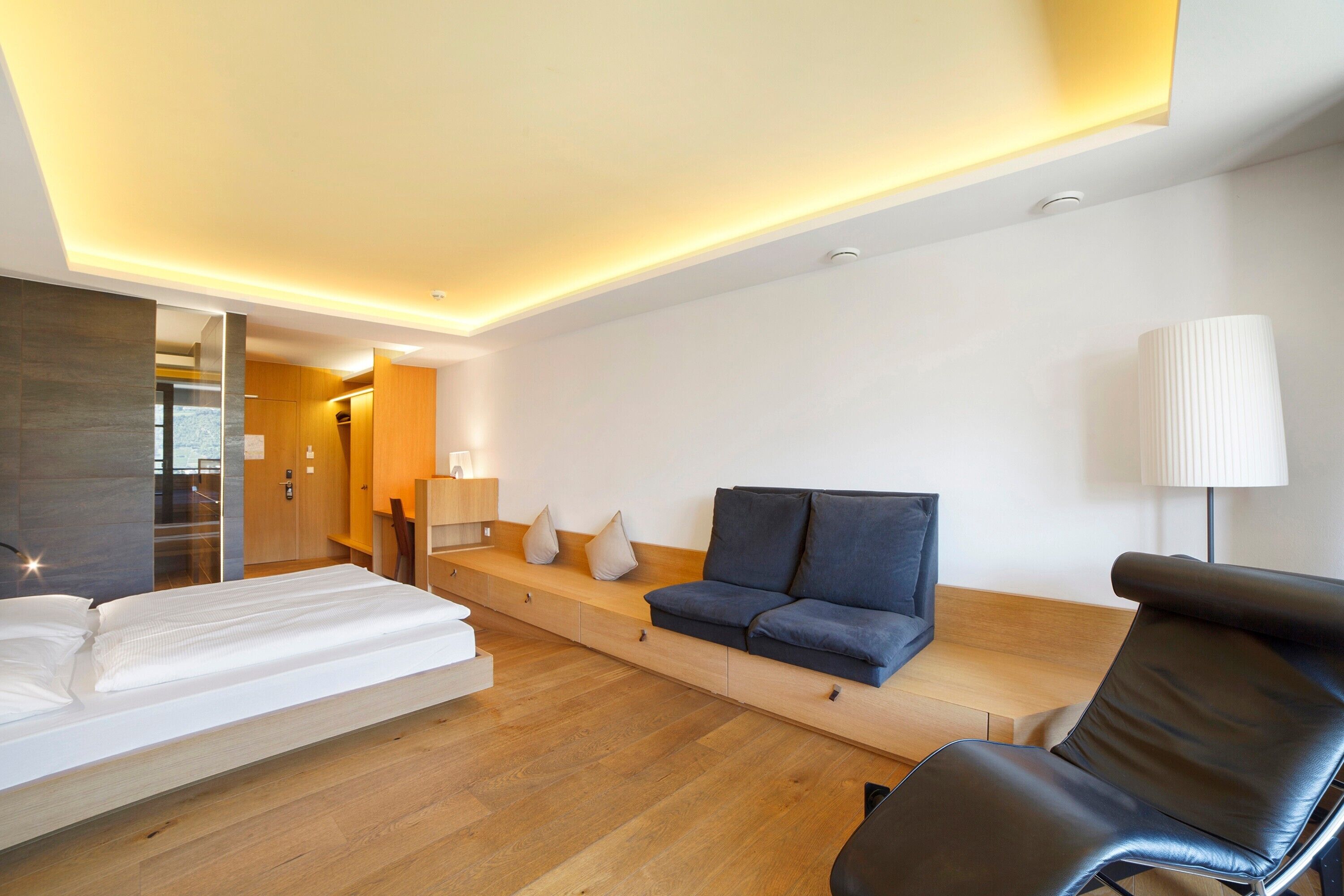 A modern hotel room with a bed, sofa, chair, and a desk with a lamp.