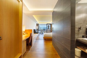 A modern hotel room with a wooden desk, a bed, a bathroom with a large mirror, and a view of the mountains.