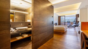 A modern hotel room with a large mirror, a built-in sink, a bed, a wooden floor, and a view of the city.