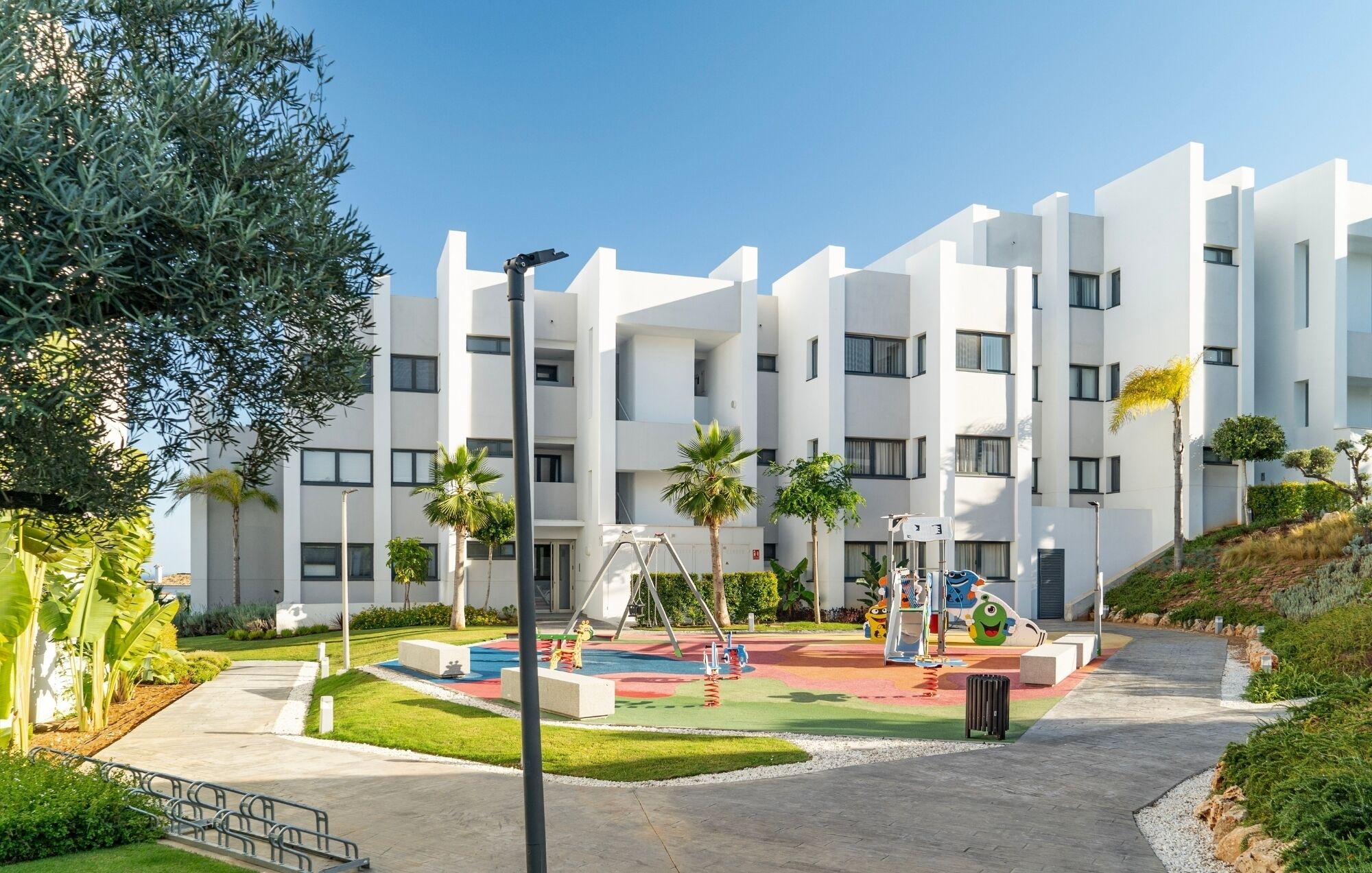Un bâtiment moderne avec une aire de jeux devant, entouré de verdure et de palmiers.