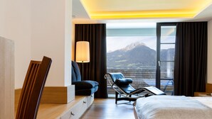 Ein modernes Hotelzimmer mit einem Bett, einem Sofa, einem Sessel, einer Lampe und einem großen Fenster mit Blick auf die Berge.