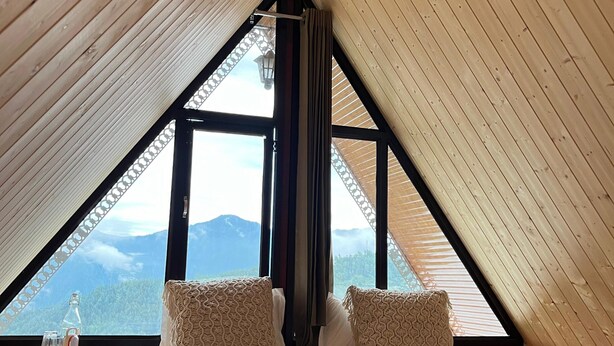 Une chambre avec un plafond en pente, une grande fenêtre et un lit agrémenté d’oreillers à motifs.