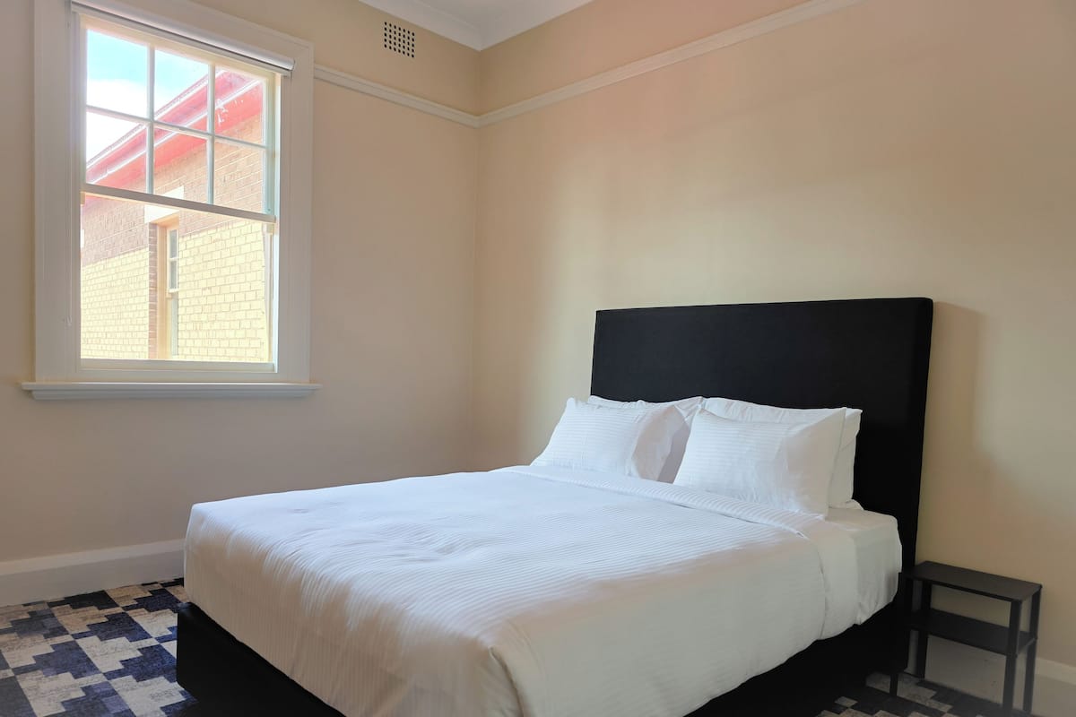 A neatly made bed with white linens in a room with a checkered floor pattern, a window with a view of a brick building, and a black headboard.