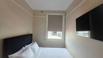 A hotel room with a bed, a ceiling fan, a window with blinds, and a TV mounted on the wall.
