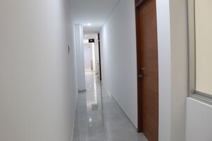 Un pasillo con una puerta de madera a la derecha, paredes blancas y suelo de baldosas.