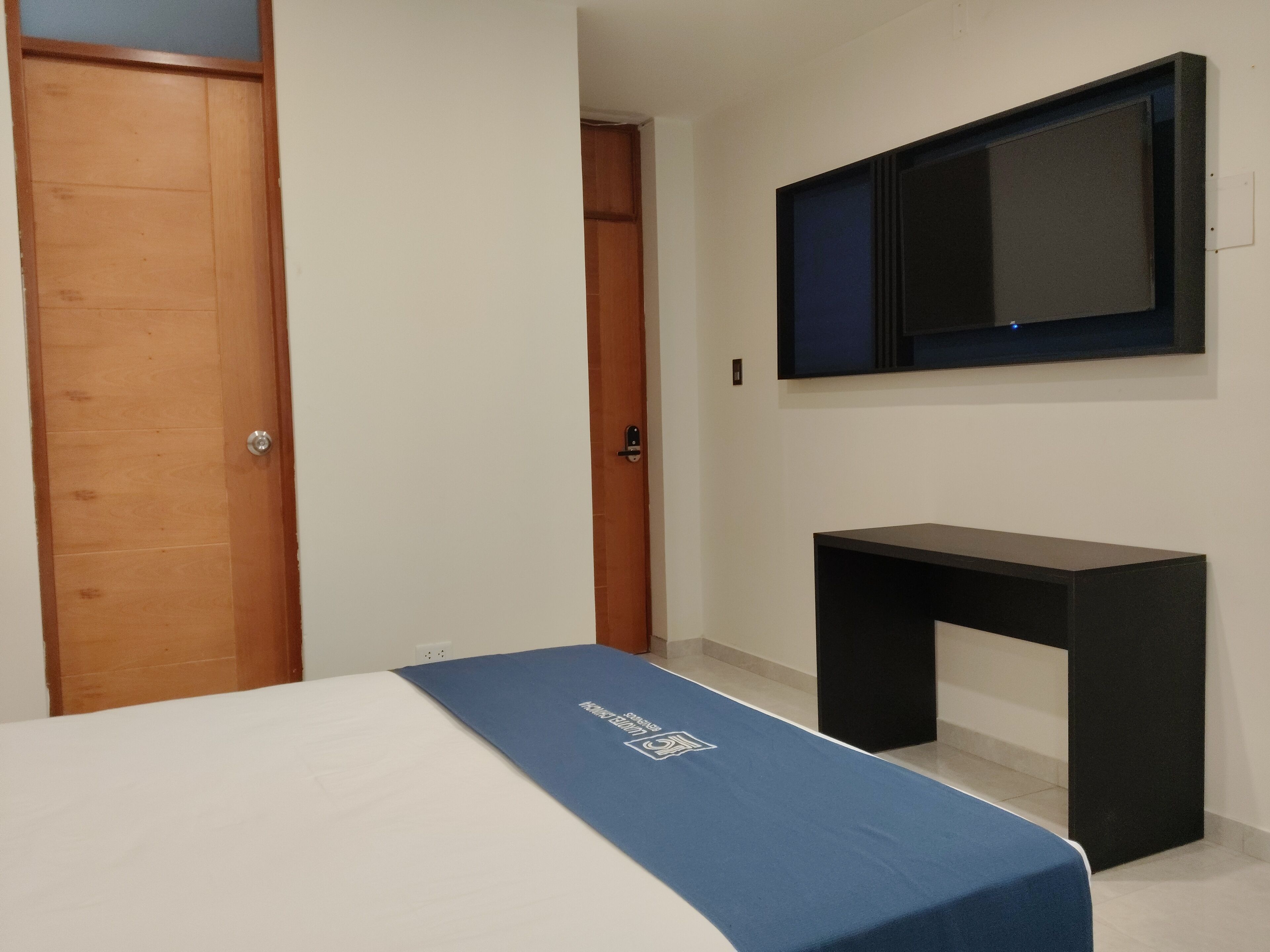 Un dormitorio con una cama, una puerta de madera, un escritorio negro y un televisor montado.