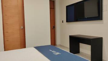 Un dormitorio con una cama, una puerta de madera, un escritorio negro y un televisor montado.