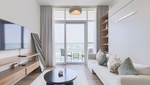 Ein modernes Wohnzimmer mit Flachbildfernseher, rundem Couchtisch, Sofa mit dekorativen Kissen, Bücherregal und großem Fenster mit Aussicht.