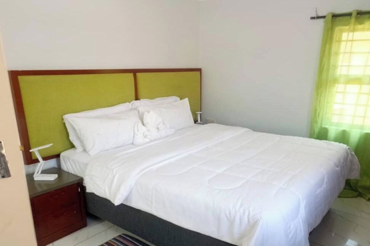 A neatly made bed with white bedding, a bedside table with a lamp, and a window with a green curtain.