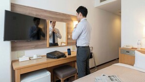 A hotel room with a bed, a desk with a chair, a mirror, a television, a phone, a water bottle, and a bedside table with a lamp.
