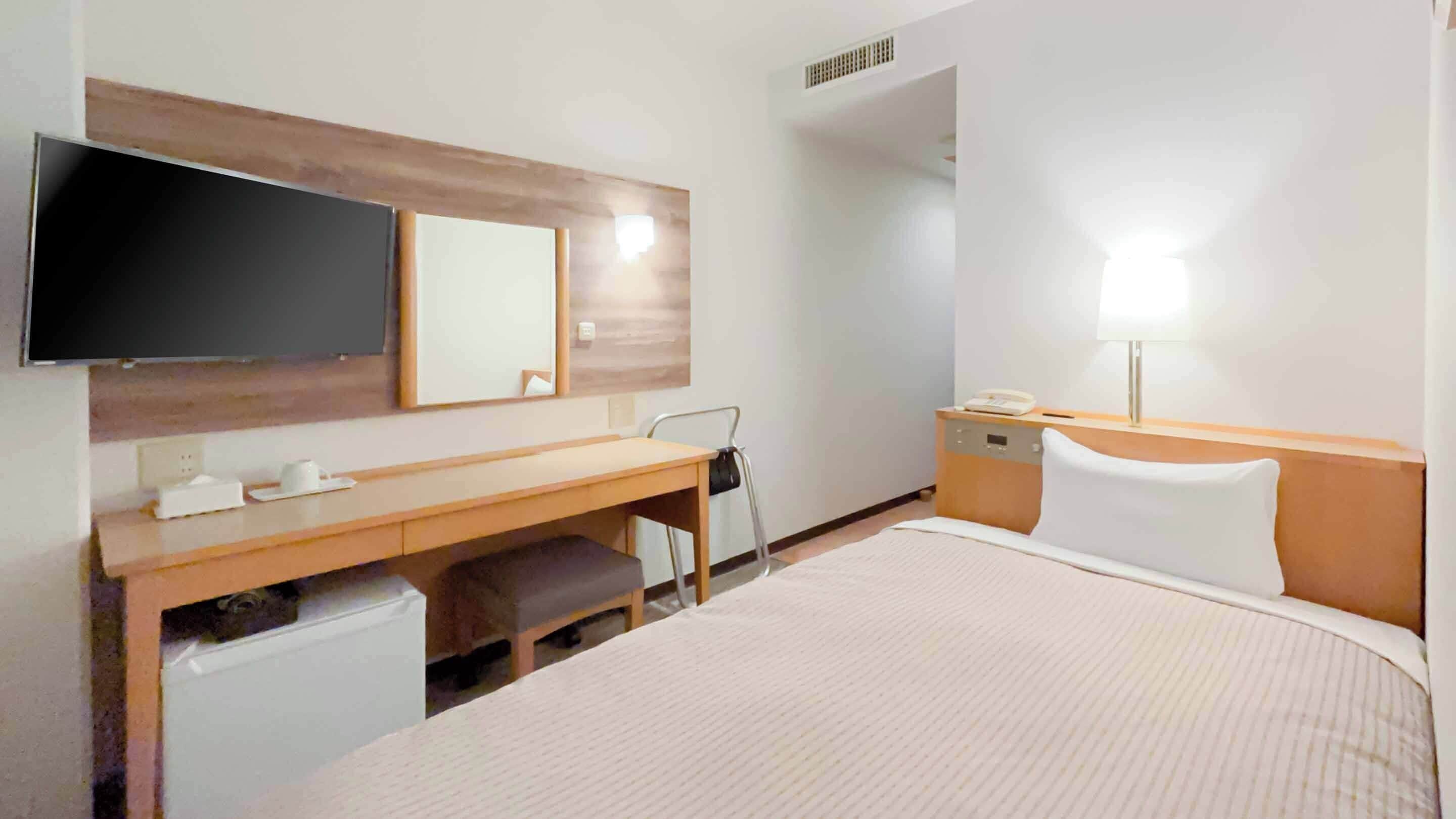 A hotel room with a bed, a desk with a chair, a television, a mirror, a bedside table with a lamp, and a small trash bin.