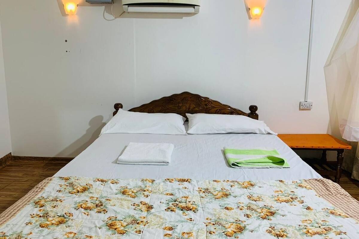 A single bed with a floral bedspread, two wall-mounted lights, an air conditioning unit, and a small table with a towel and green cloths.