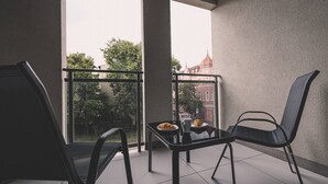 Un balcón con dos sillas, una mesa, una barandilla de cristal y vista a un edificio con tejado rojo.