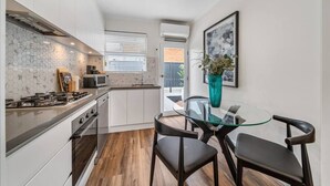 Una cocina moderna con armarios blancos, una estufa a gas y un microondas. Hay una mesa de vidrio con dos sillas. Un jarrón con flores está sobre la mesa.