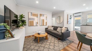 A modern living room with a grey sofa, a round coffee table, and a green armchair. There are abstract paintings on the wall and a large mirror above the sofa. - One Tea House—Contemporary Cityside Comfort (Mile End)