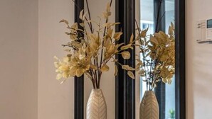 Un interior moderno con un espejo de marco negro, dos jarrones blancos con plantas secas y un pequeño objeto negro sobre una consola oscura.