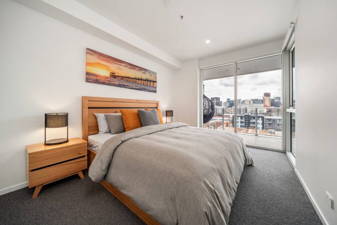 Une chambre à coucher moderne avec un grand lit, une table de chevet, une lampe, une vue sur la ville à travers une grande fenêtre et un tableau représentant un coucher de soleil accroché au mur.