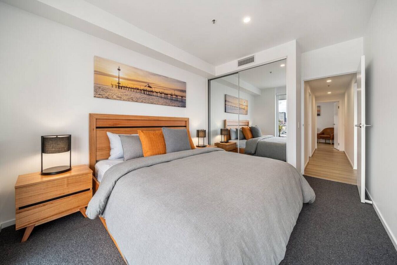 A modern bedroom with a large bed, wooden headboard, and a nightstand with a lamp. A painting of a beach scene is on the wall, and there is a mirror reflecting the room.