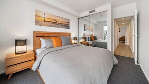 Une chambre moderne avec un grand lit, une tête de lit en bois et une table de chevet avec une lampe. Un tableau représentant une scène de plage est accroché au mur, et un miroir reflète la pièce.