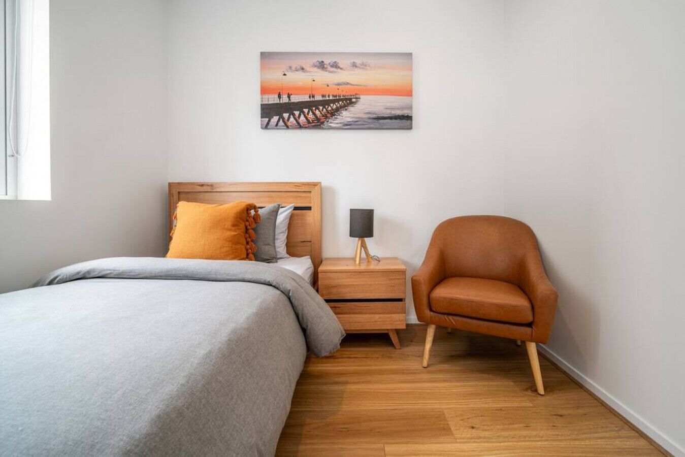 Een slaapkamer met een bed, een nachtkastje, een stoel en een aan de muur hangend schilderij van een zonsondergang boven een pier.