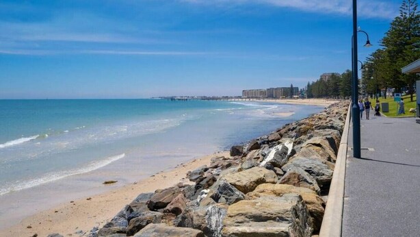 A coastal walkway with a rocky barrier, a clear blue sky, and a distant pier. - Salt at Arlo`s - Cosy Comfort near Glenelg Beach (Glenelg South)