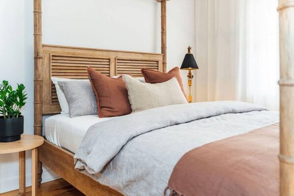 Un lit bien fait, avec une tête de lit en bois, une table de chevet ornée d’une plante et une lampe.