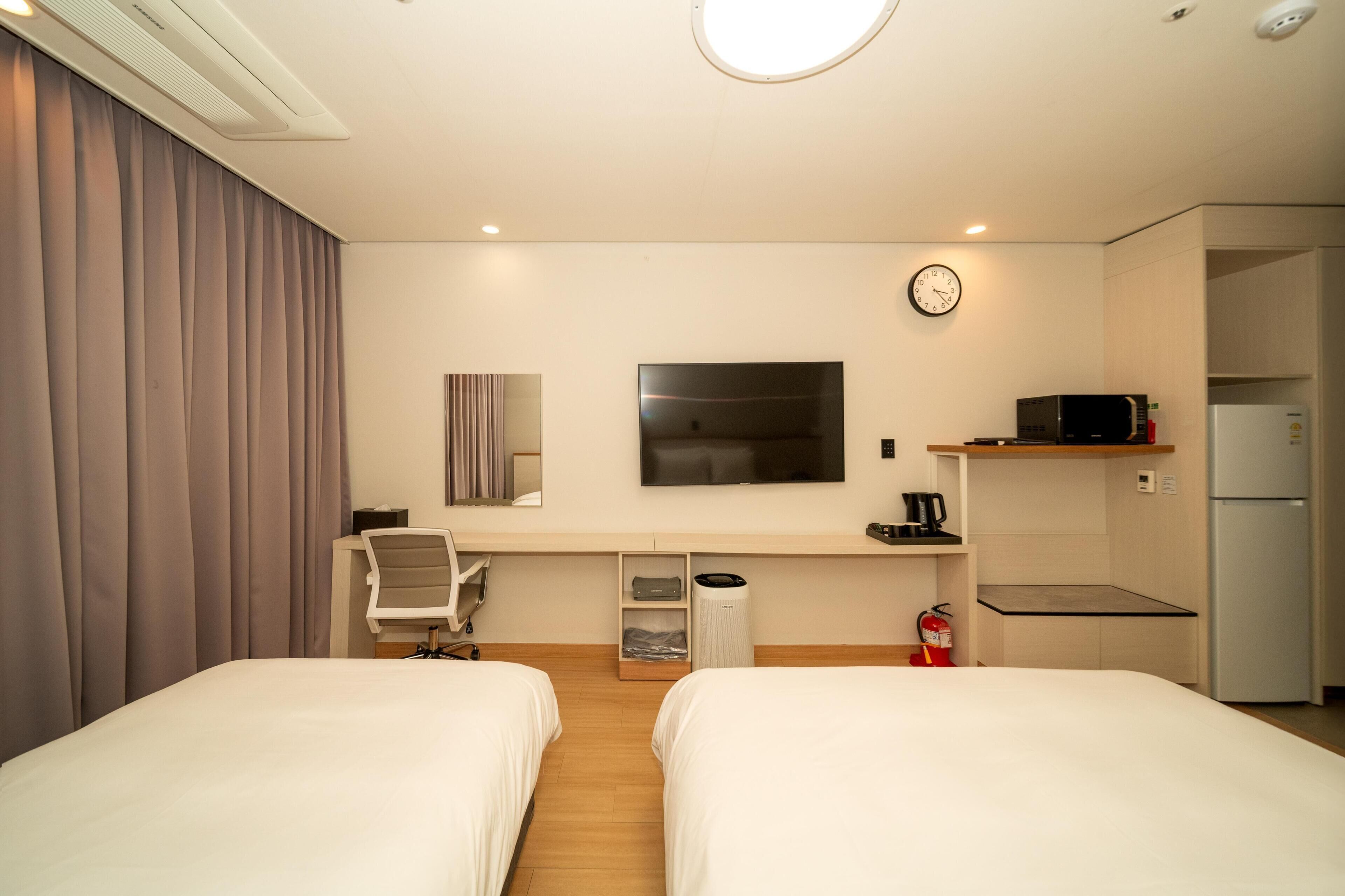 A hotel room with two beds, a desk, a chair, a television, a clock, a microwave, and a refrigerator.