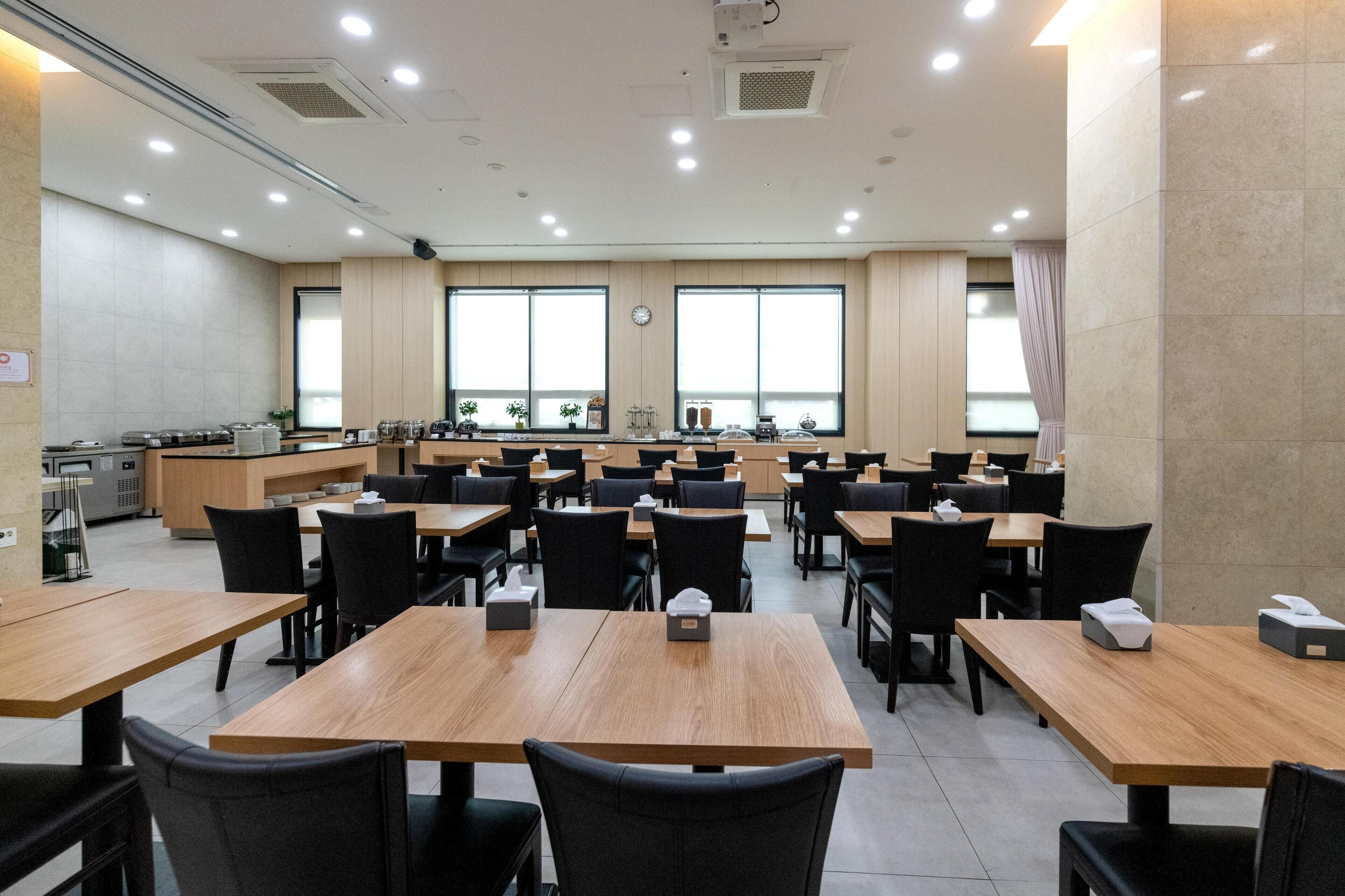 A spacious dining area with multiple tables and chairs, a kitchen area, and large windows providing natural light.