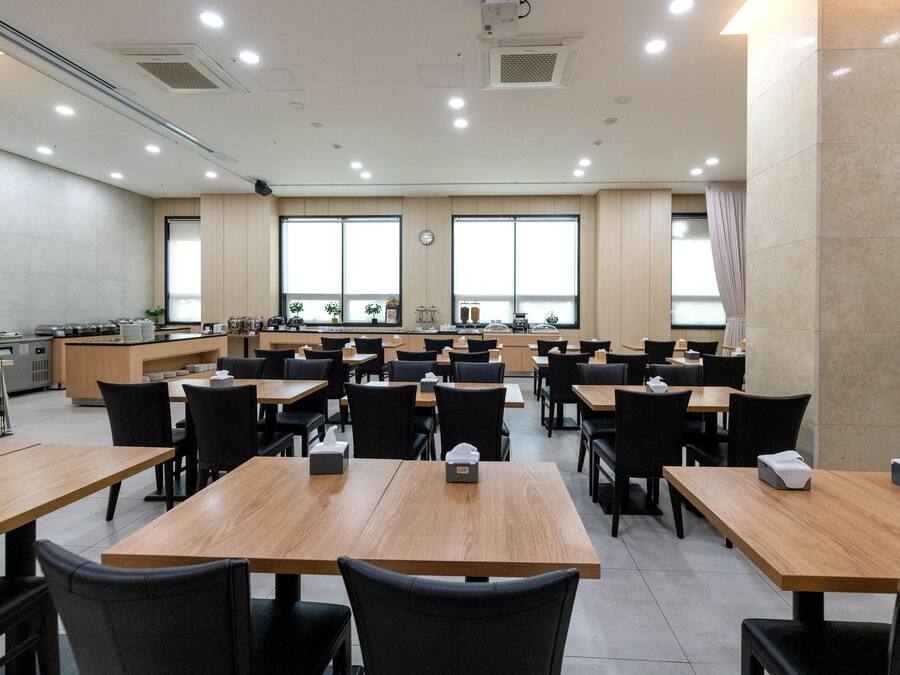 A spacious dining area with multiple tables and chairs, a kitchen area, and large windows providing natural light.