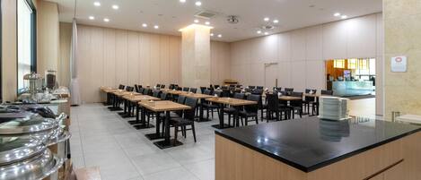 Un salón de conferencias con varias mesas y sillas, una zona de buffet con recipientes de comida y un mostrador de recepción.