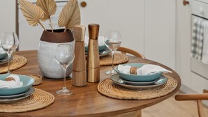 Une table de salle à manger ronde en bois, avec de l’assiette et des verres bleus, entourée de sets de table en fibres tressées.
