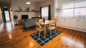 Moderni keittiö, jossa on valkoiset kaapit, ruokailutila puupöydällä ja -tuoleilla, harmaa sohva ja tuuletin.