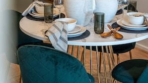 A dining area with a round table set for four, featuring white plates, bowls, and glasses. There are two large white vases and a small green vase on the table. The chairs are teal with gold legs. - Paradiso on Bent—City Chic near Rundle Mall (Adelaide)