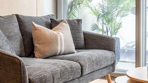 A modern living room with a grey sofa, wooden shelves, a round wooden table, and a large window with a view of buildings and a plant. - Paradiso on Bent—City Chic near Rundle Mall (Adelaide)