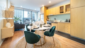 A modern kitchen with wooden cabinets, a dining area with a round table and chairs, and a view of the city through large windows. - Paradiso on Bent—City Chic near Rundle Mall (Adelaide)