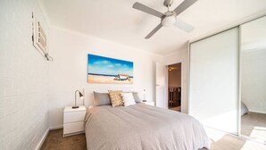 Une chambre à coucher comprenant un lit, une table de chevet, un ventilateur de plafond, un tableau représentant une scène de plage et une porte-fenêtre coulissante donnant sur un balcon.