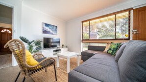 Un salon avec un canapé, un téléviseur, une table basse blanche et un fauteuil en osier.