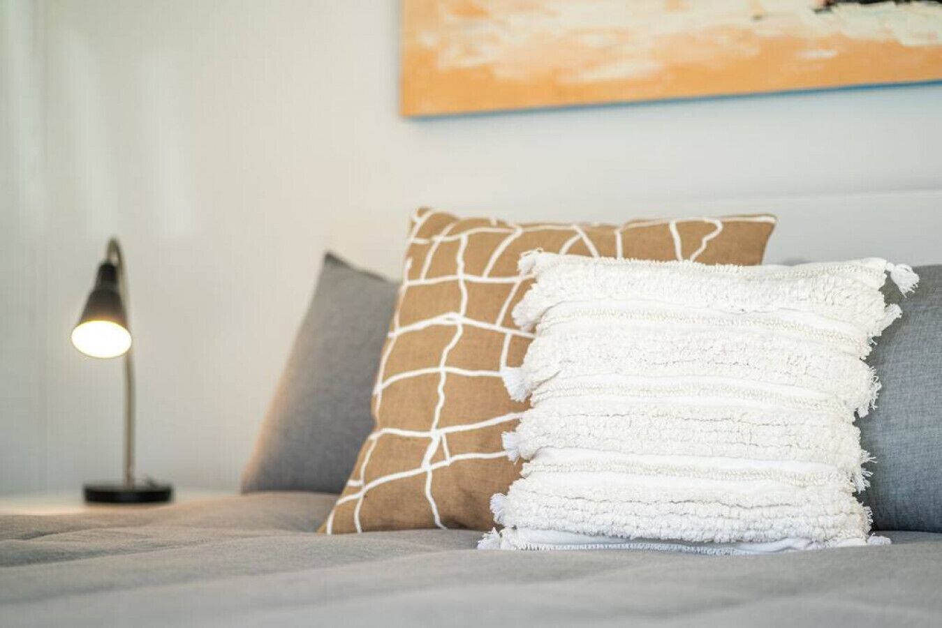 Una cama con un edredón gris, una almohada blanca texturizada y una almohada marrón con un diseño blanco. Se ve una lámpara de pie con pantalla blanca al fondo.
