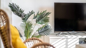 Un salon moderne avec un téléviseur à écran plat, un fauteuil en osier doté d’un coussin jaune et une grande plante verte.