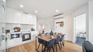 A modern kitchen with white cabinets, built-in ovens, and a dining area with a wooden table and black chairs. - In The Midst on Melbourne - Walk to Cafés & Dining (North Adelaide)