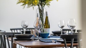 Mesa lista para comer con platos, tazones y copas de vino, con una botella de vino y un florero con flores amarillas al fondo.