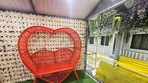 A red heart-shaped chair in a room with a wall covered in white flowers, a yellow bench, and a view of a garden through a glass wall. - Astha Home Shared Pool Rooms (Mumbai)