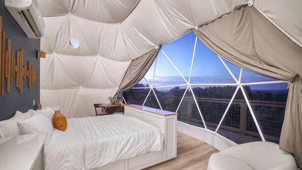 A bedroom with a bed, a desk, and a chair. There is a large window with a view of a bridge and trees. - Luxury Dome - Spa Shower - Panoramic Views (Spring Branch)