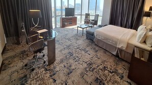 Quarto de hotel com uma cama grande, uma escrivaninha, uma cadeira, uma televisão e vista da cidade através de amplas janelas.