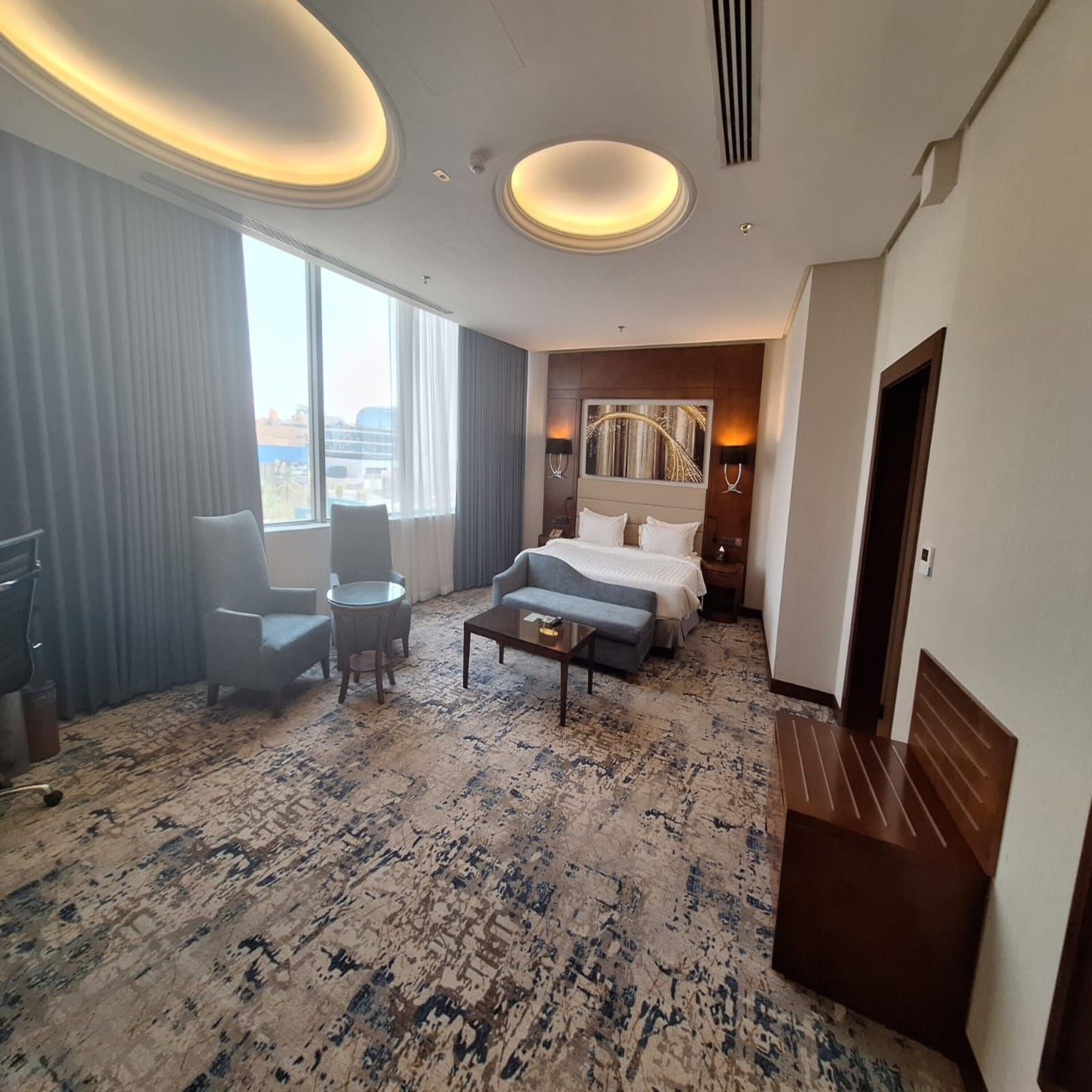 A hotel room with a bed, a desk, a chair, a coffee table, and a view of the city through large windows.