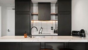 Una cucina moderna con mobili scuri, un piano di lavoro bianco e un piano cottura integrato. Ci sono due sgabelli all'isola e una macchina da caffè sul bancone.