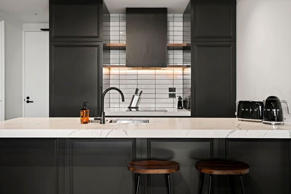 A modern kitchen with dark cabinetry, a white countertop, and a built-in stove. There are two stools at the island and a coffee maker on the counter.