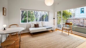A modern living room with a sofa, a dining table, and chairs. Large windows provide natural light and views of the outdoors.