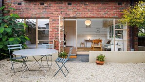 A brick house with a patio area featuring a round table and chairs, a small garden with plants, and a sliding glass door leading to an interior space with a sofa and a table.