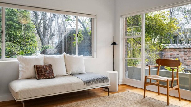 A cozy room with a sofa, a chair, and large windows offering a view of greenery and a brick wall.